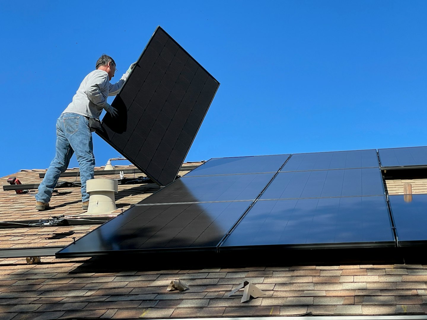 Un technicien installe un panneau solaire sur un toit en bardeaux sous un ciel bleu.